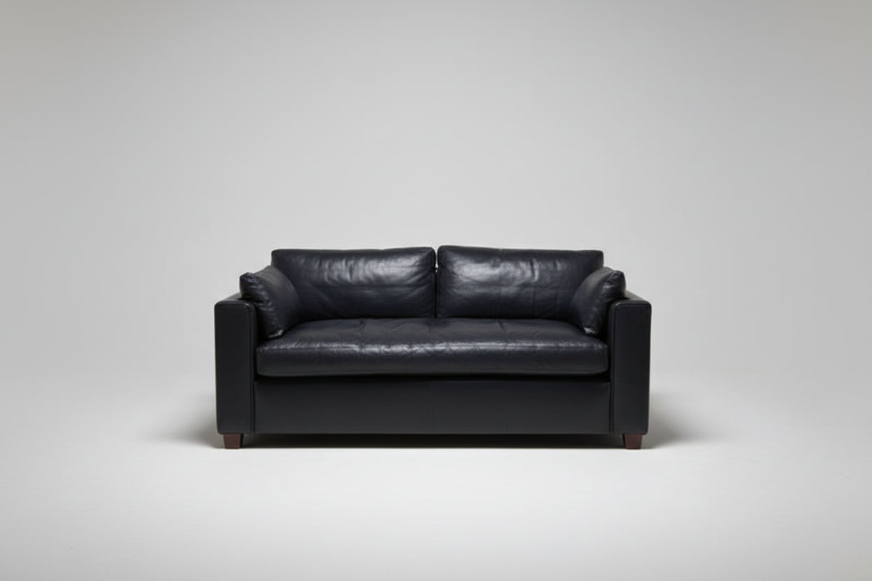 A black leather loveseat sofa with two back cushions and matching armrests, photographed in a room with a light-colored wall, carpet, a side table with a lamp on the left, and another lamp on the right.