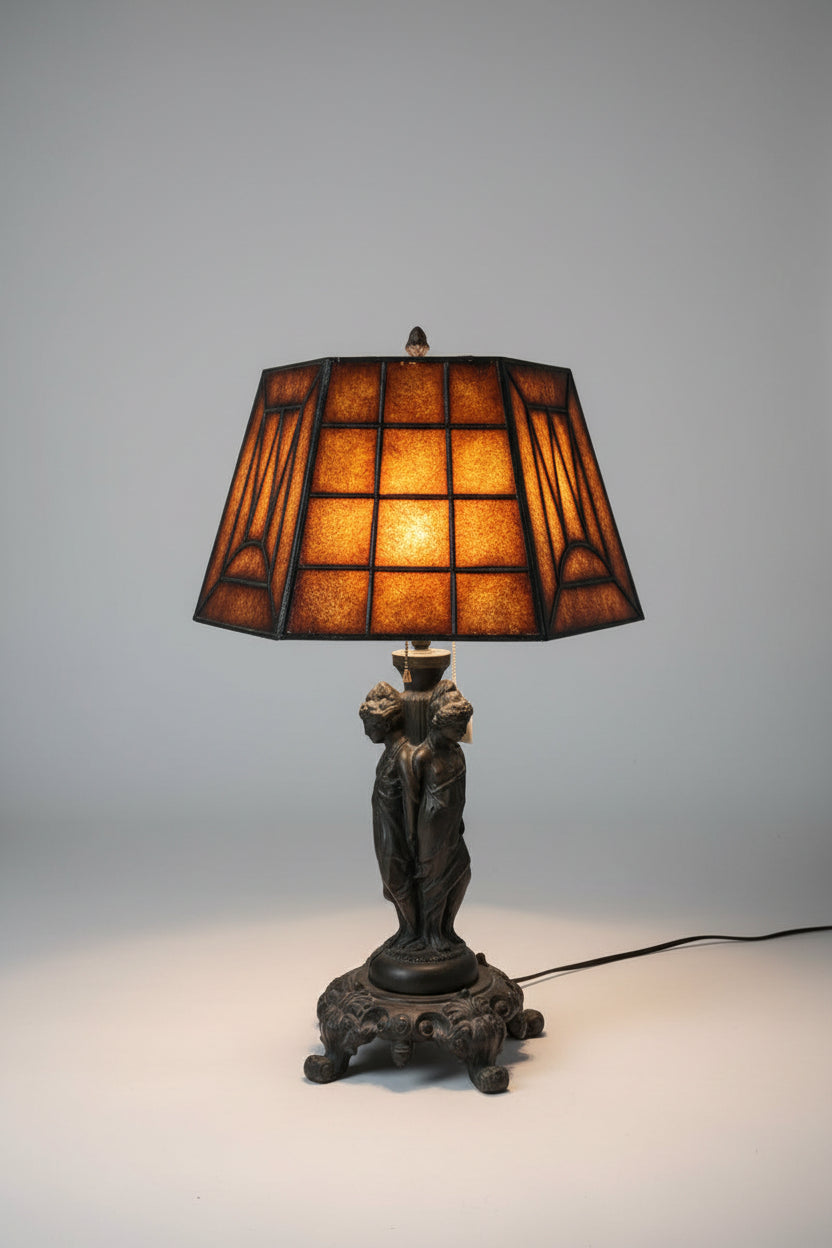 A vintage-style table lamp with a warm amber stained-glass shade and a dark sculptural base, displayed on a wooden shelf in a cluttered antique shop or home decor store alongside bowls, books, and framed art.