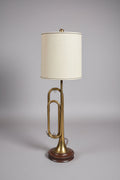 A brass trumpet-shaped table lamp with a beige fabric shade and a decorative black base, photographed in a cluttered indoor room with furniture and pillows in the background.