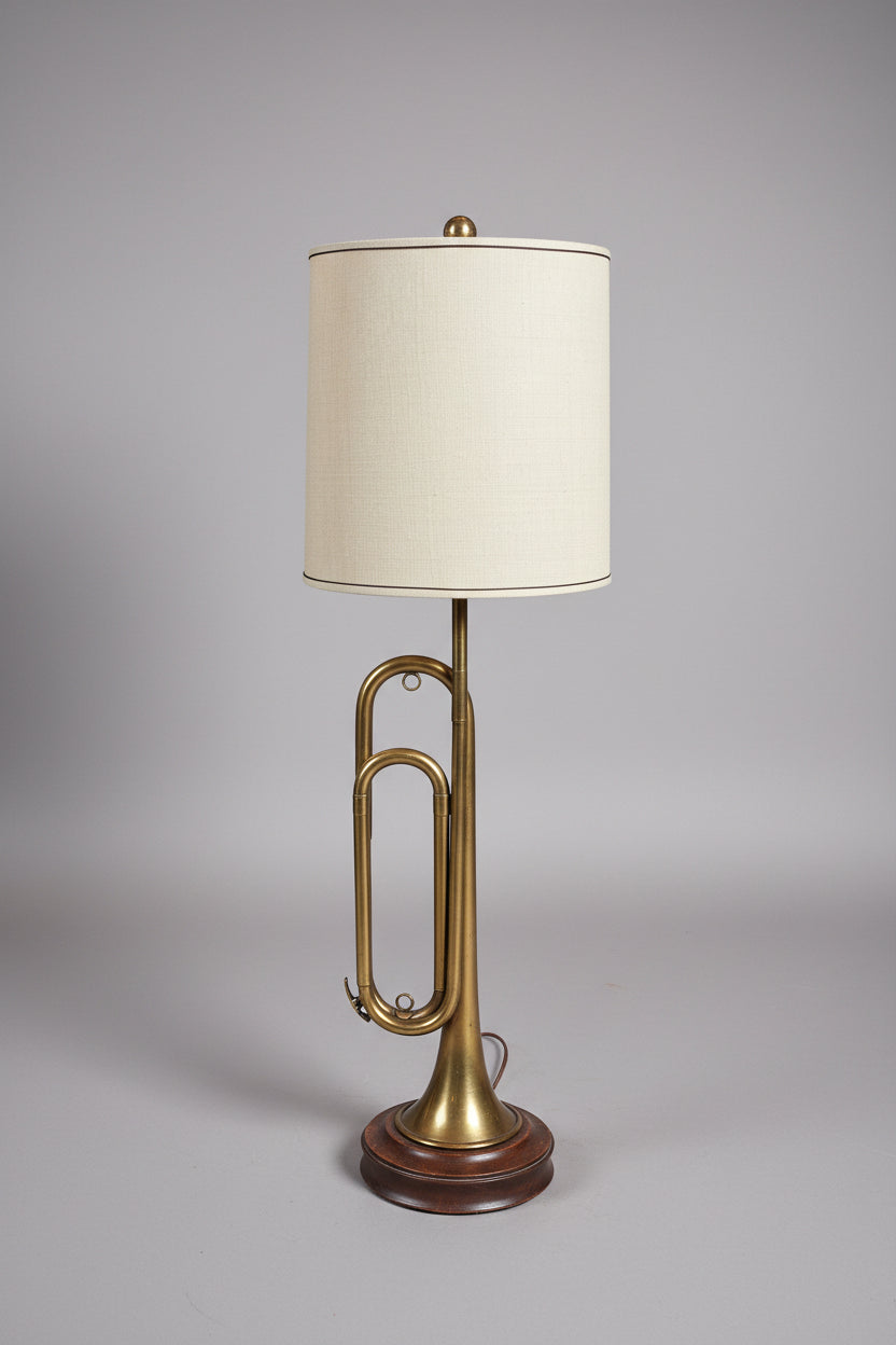 A brass trumpet-shaped table lamp with a beige fabric shade and a decorative black base, photographed in a cluttered indoor room with furniture and pillows in the background.
